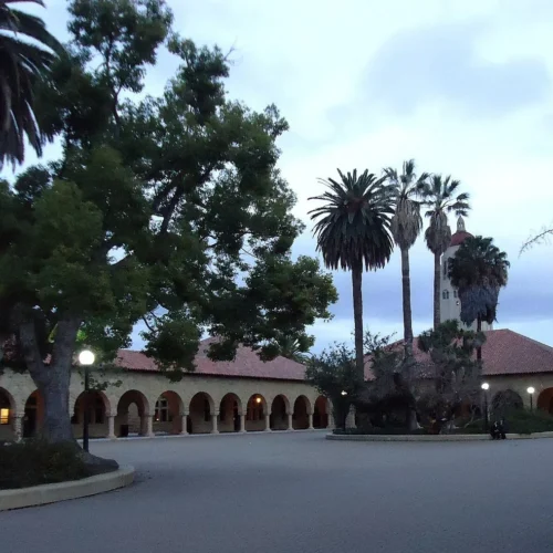 Arched university buildings and palm trees on the Stanford campus at twilight.