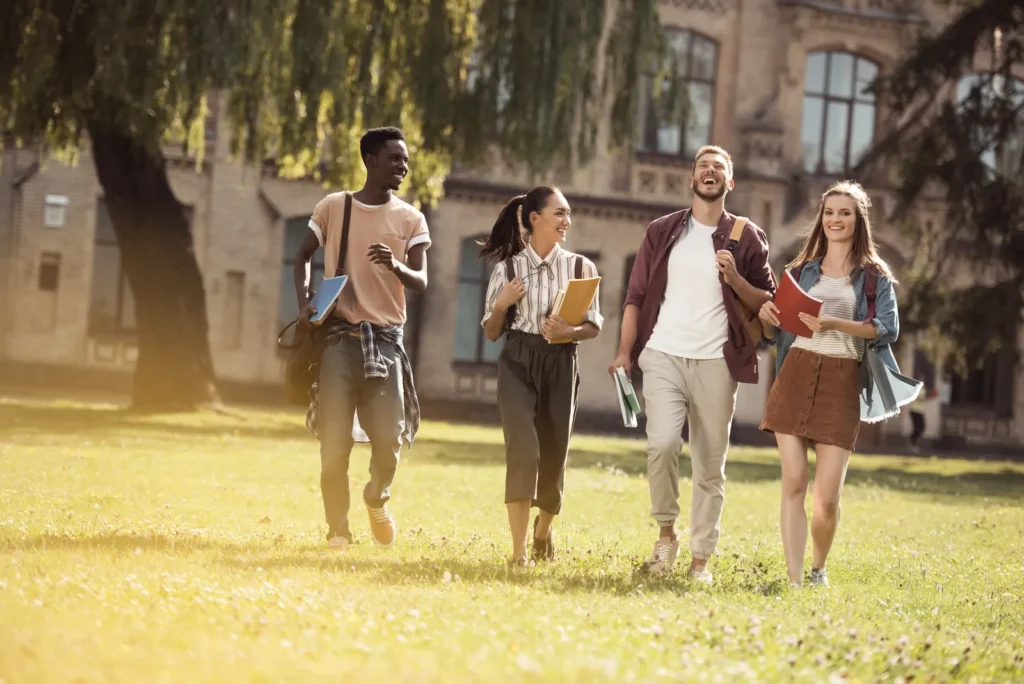 Four young adults carrying books across a sunlit university lawn.