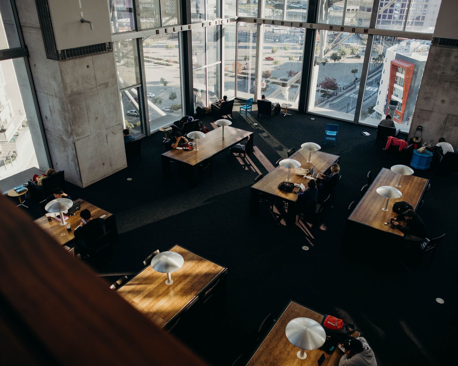 A high-angle view of people studying at tables beside large library windows.